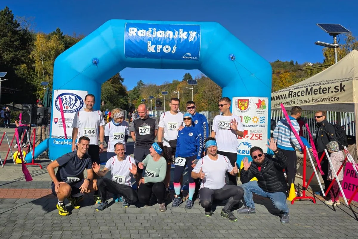 Veľká skupina bežcov v športovom oblečení pózuje pred modrým nafukovacím štartovacím oblúkom na preteku.