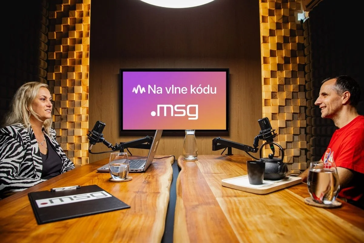 Žena a muž sedia oproti sebe v štúdiu s mikrofónmi, za nimi svieti obrazovka s názvom podcastu Na vlne kódu.